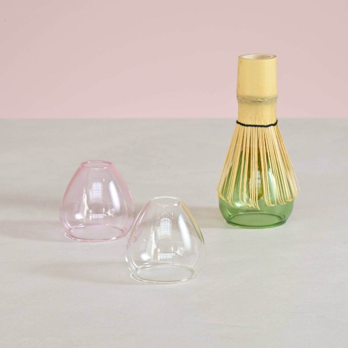 Three glass whisk stands in a pink, clear, and green base on a light gray surface with a pink background.
