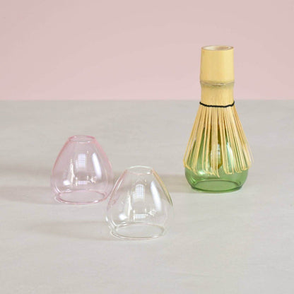 Three glass whisk stands in a pink, clear, and green base on a light gray surface with a pink background.