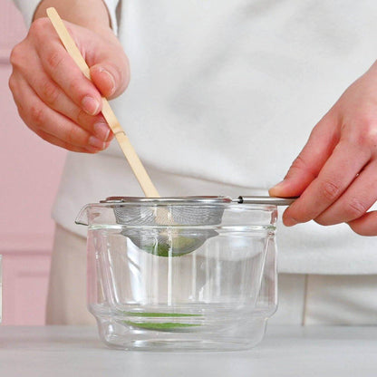 Person using a bamboo scoop and a strainer over a glass bowl on a white background