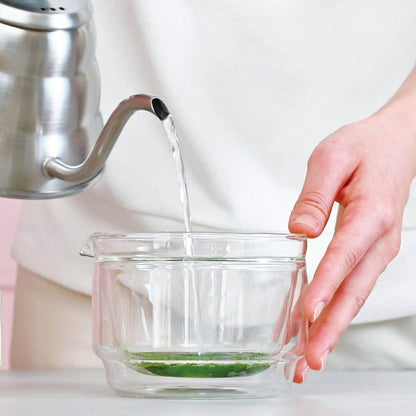 Person pouring water from a silver kettle into a clear glass bowl with matcha on a white background