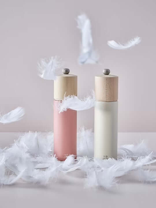 Two salt grinders in white and dark pink with wooden tops on a surface with feathers